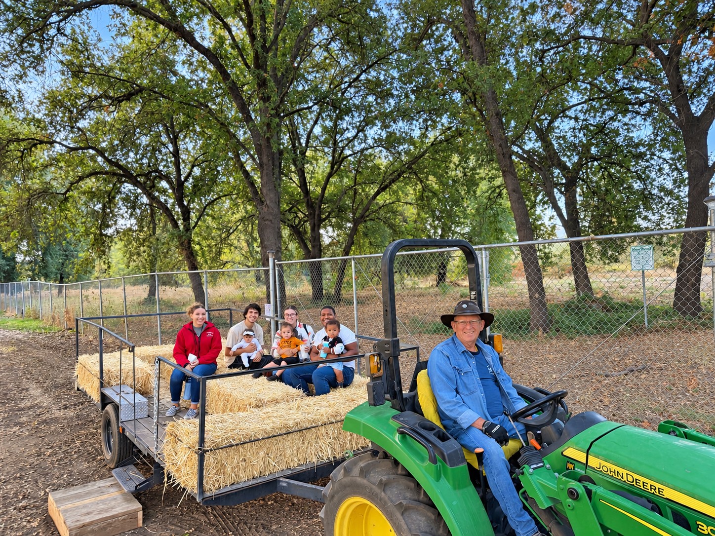 Children and adults sitting on hay bales in a farm trailer beside a tractor and its smiling driver